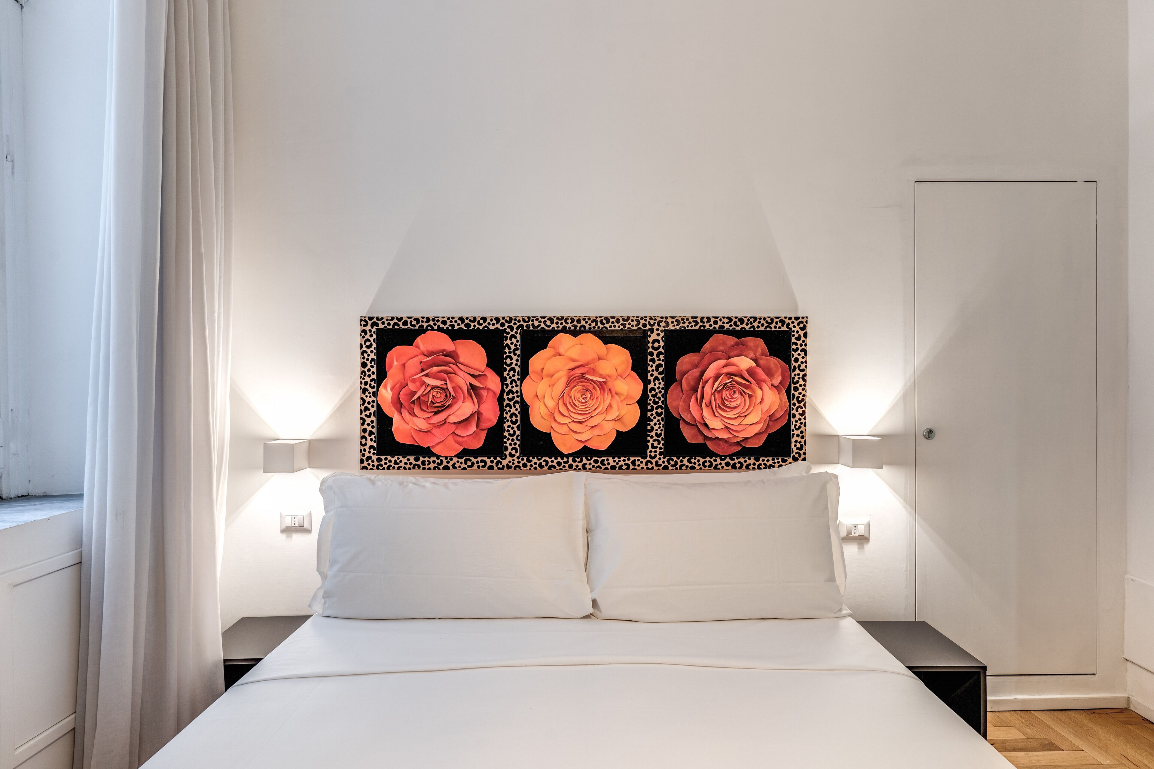A bed with white linens, two bedside tables, and a wall-mounted artwork of three large roses.