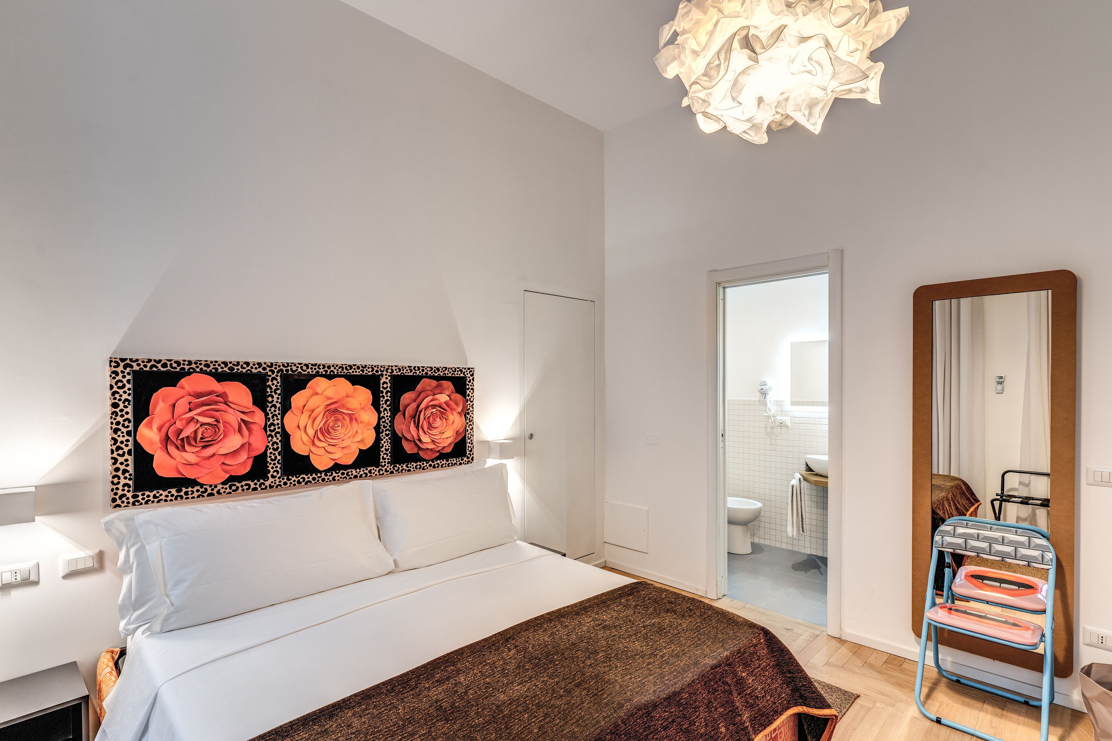 A bedroom with a bed, a wall-mounted artwork of roses, a mirror, and a bathroom visible through an open door.
