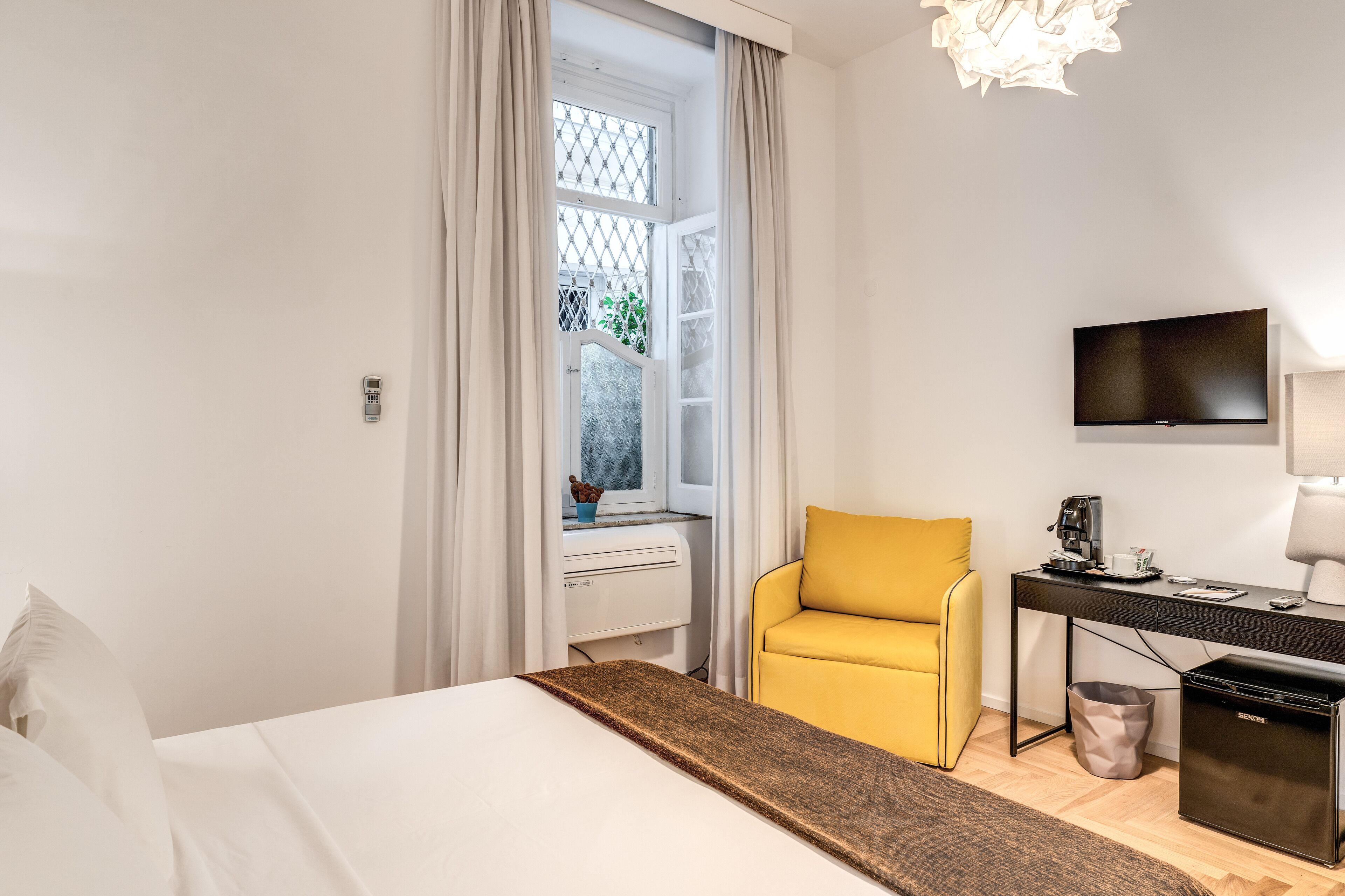 Une chambre d’hôtel avec un lit, un fauteuil jaune, un bureau avec un ordinateur, une télévision et une fenêtre avec des rideaux.