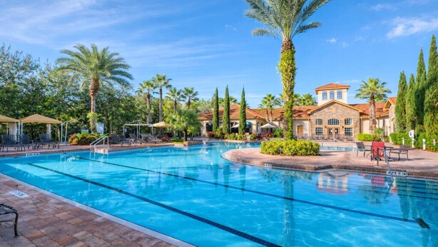 A swimming pool with lounge chairs and umbrellas, surrounded by palm trees and a building with a tiled roof. - 1301 Flamingo Cove at Tuscana (Davenport)