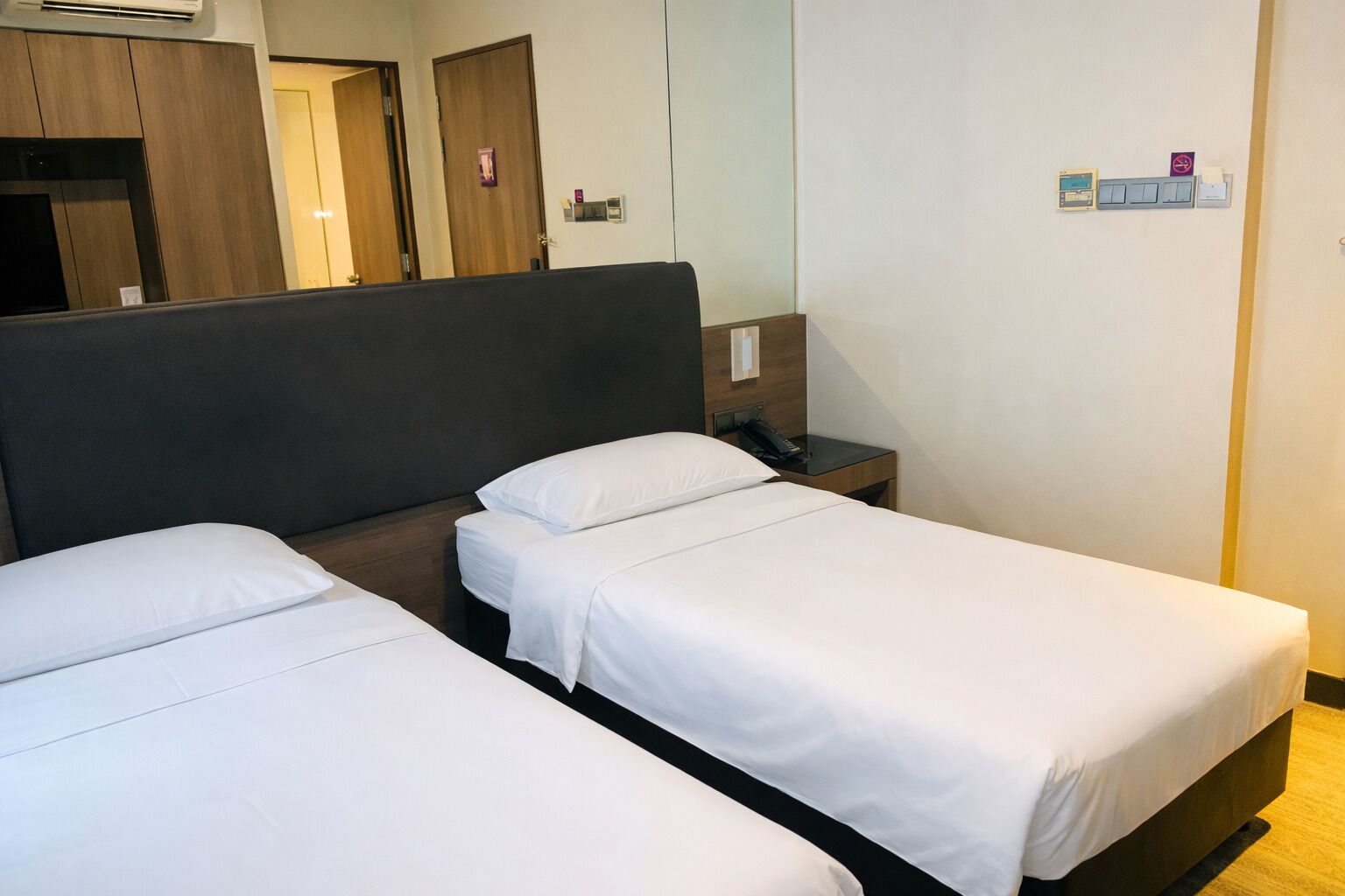 A hotel room with two single beds, a wooden headboard, a bedside table with a phone, and a wall-mounted light switch.