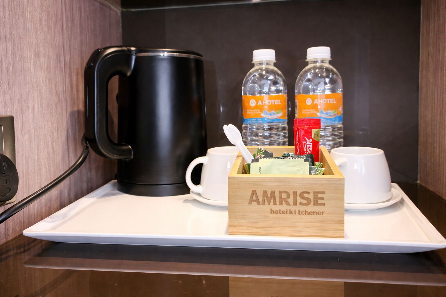 Eine Hotelküche mit einer schwarzen Kaffeemaschine, zwei Wasserflaschen, einer mit "AMRISE Hotelküche" beschrifteten Schachtel und zwei weißen Tassen auf einem Tablett.