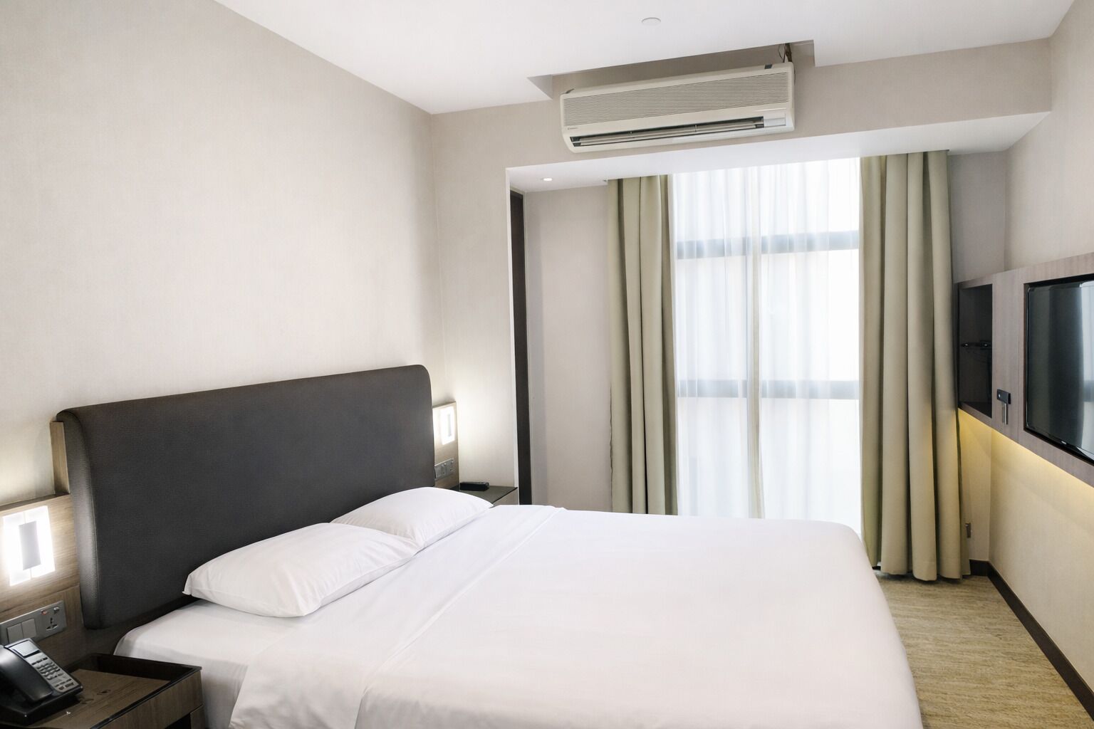 A hotel room with a large bed, an air conditioning unit, a TV, and a window with curtains.