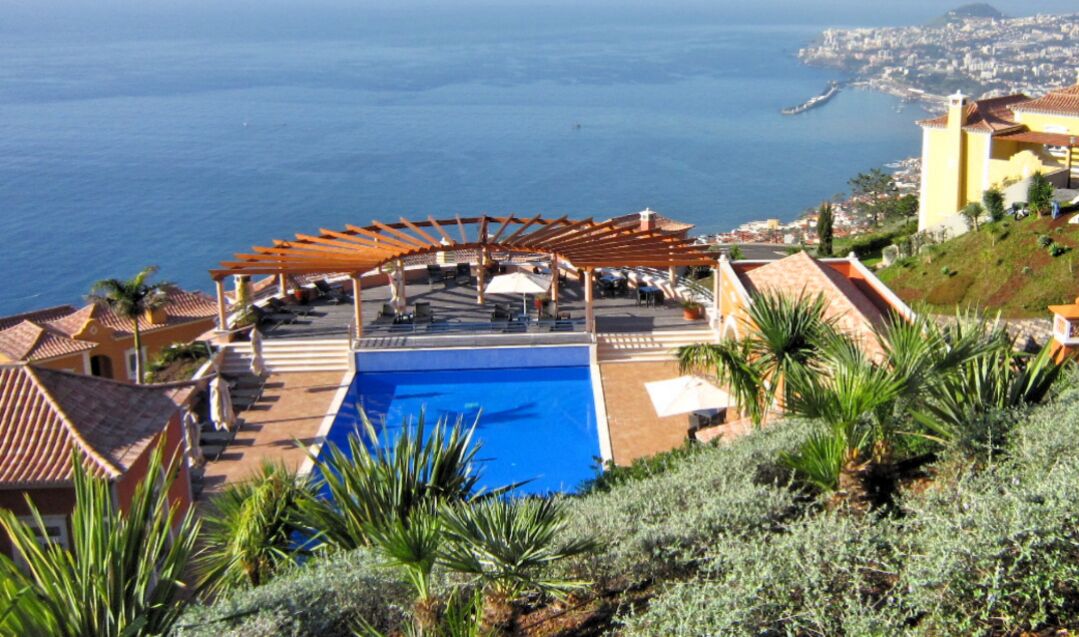 A pool area with a pergola, lounge chairs, and a view of the sea.