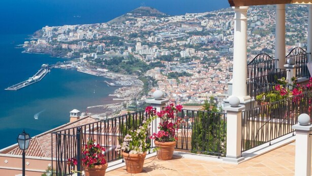 Un balcon orné de pots en terre cuite remplis de fleurs, une vue sur le paysage urbain de la côte et une mer bleue et limpide.