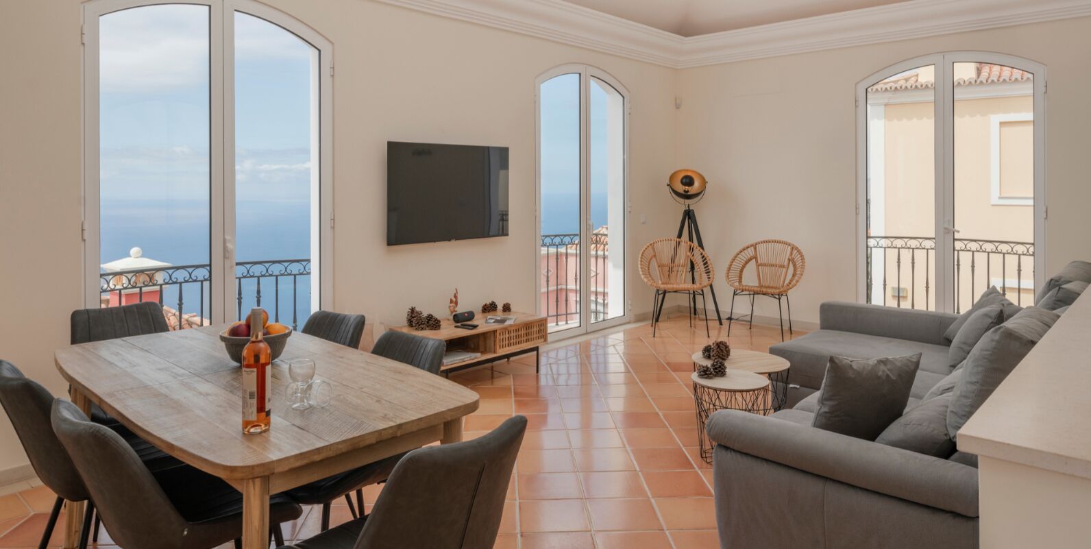 A spacious living area with a wooden dining table, grey sofa, and wicker chairs. A flat-screen TV is mounted on the wall, and there are large windows offering a view of the sea.