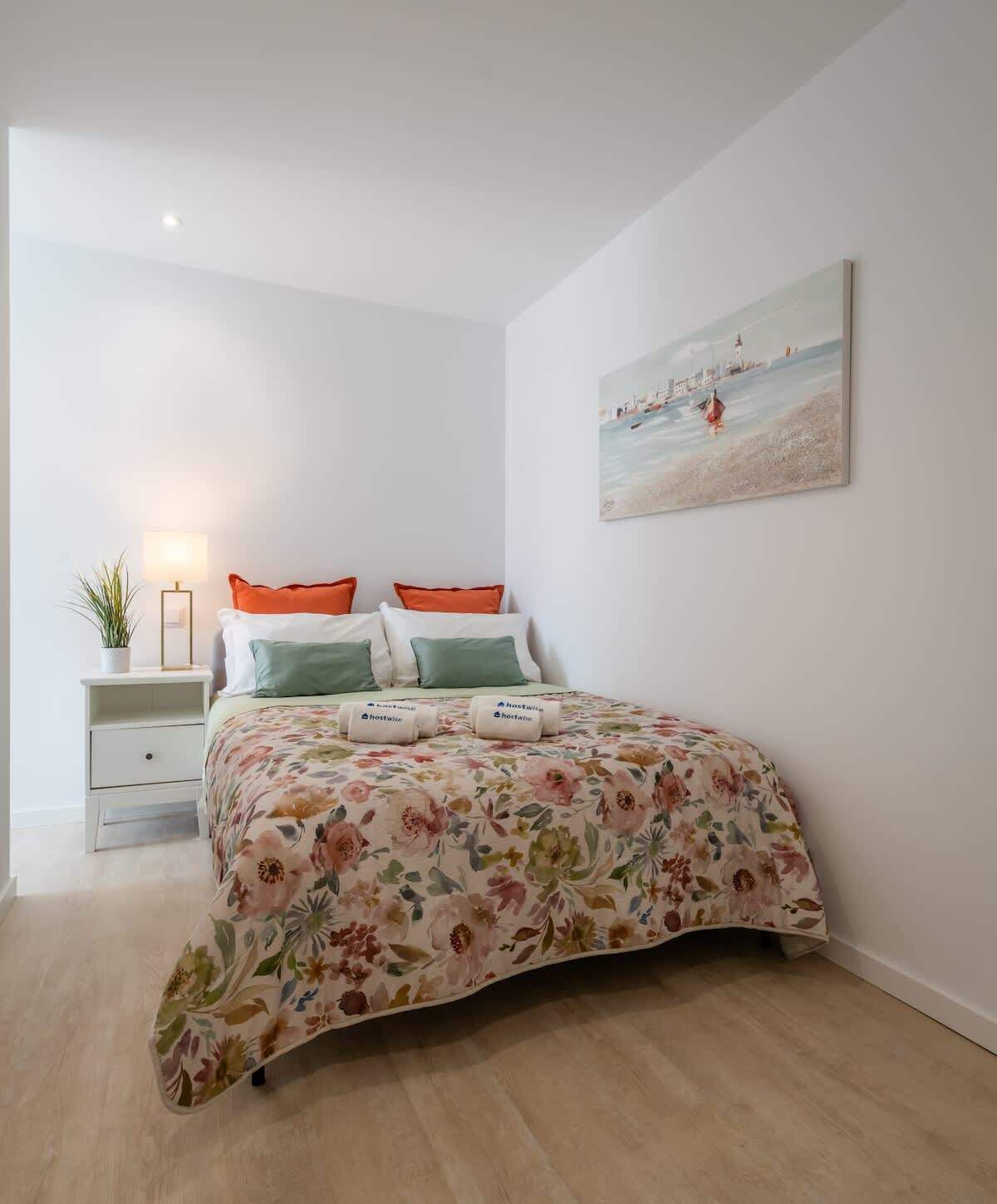 Une chambre à coucher avec une couvre-lit à motifs floraux, une table de chevet avec une lampe et un tableau représentant une scène de plage.