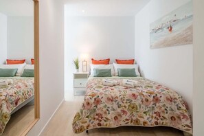 Un dormitorio con dos camas, cada una con ropa de cama floral y almohadas naranjas, una mesita de noche con lámpara, y un cuadro en la pared.