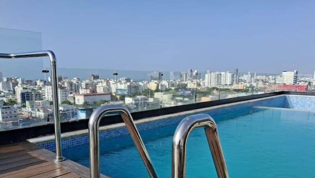 Una piscina en la azotea con barandillas metálicas y una terraza de madera, con vistas a un paisaje urbano.