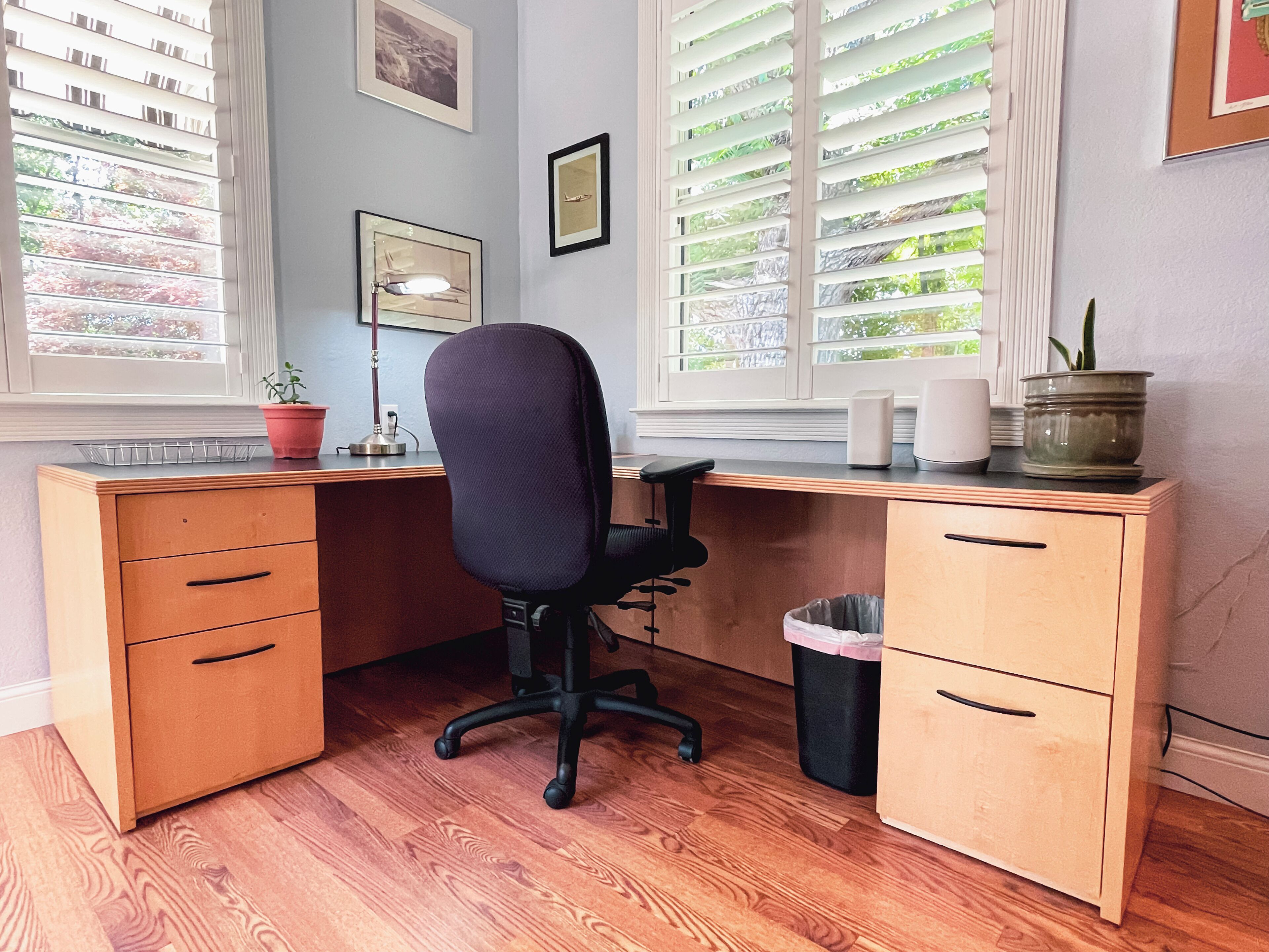 A wooden desk with a black office chair, a potted plant, a lamp, and a trash can.