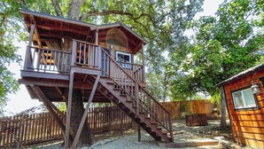 Une cabane en bois perchée dans un arbre, accessible par un escalier, et entourée d’arbres et d’une clôture en bois.