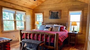 A cozy cabin bedroom with a wooden bed, a bear-themed pillow, and a window with a view of trees. - Stunning Views From Lodge, and With hot Tub! (Blue Ridge)