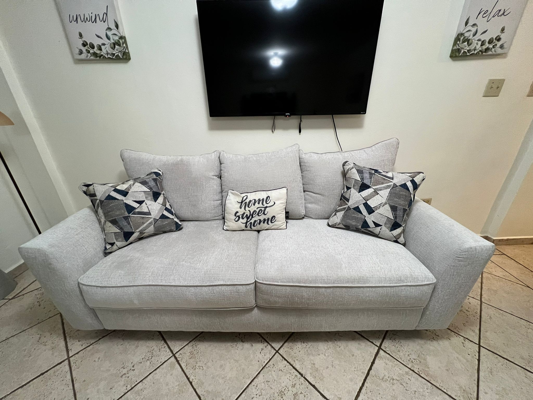 Ein graues Sofa mit gemusterten Kissen, ein Flachbildfernseher an der Wand und zwei Wanddekorationen mit Text.