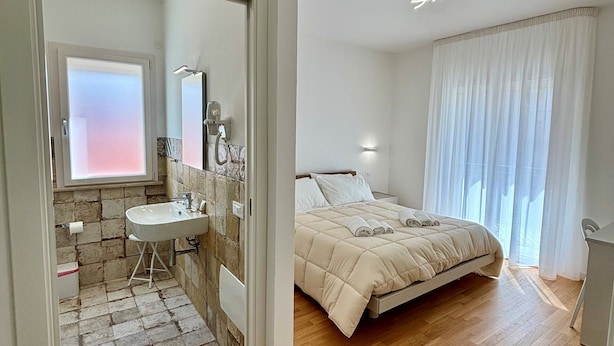 Una camera da letto con un letto, un comodino, una finestra con tende e un bagno con lavandino e specchio.