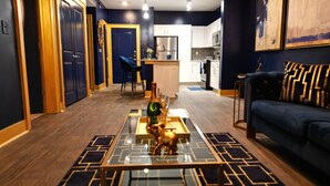 A modern living room with a glass coffee table, a blue sofa, and a kitchen in the background. - Live Like ROYALTY in Buckhead This unique place has a style all of its own. (Atlanta)