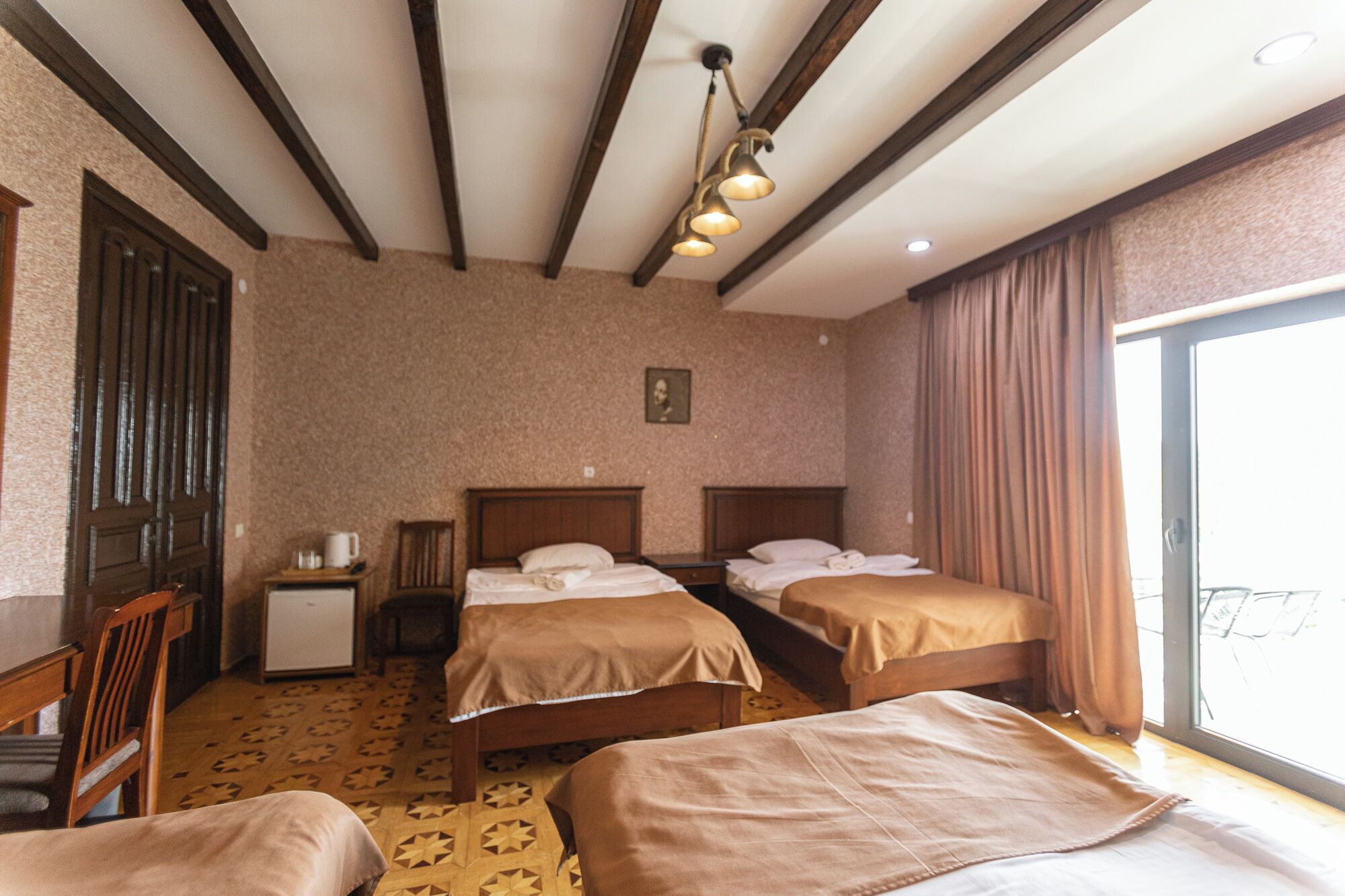 A hotel room with two single beds, a small desk, a chair, a wardrobe, and a large window with curtains.