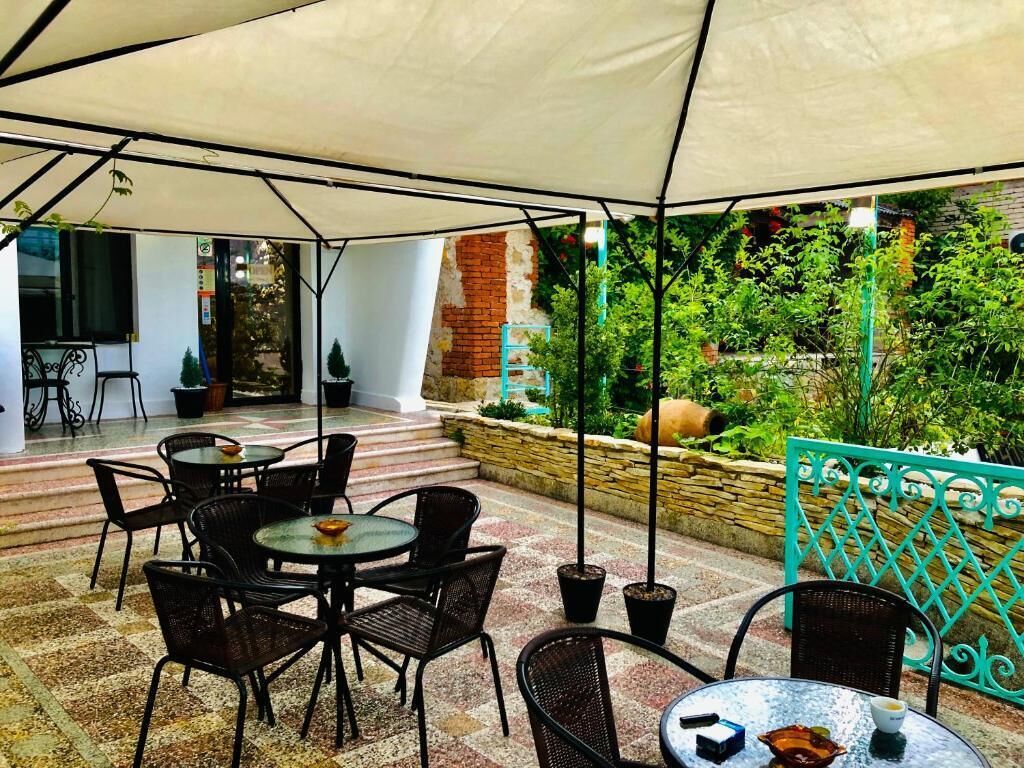 Un patio al aire libre con mesas y sillas redondas, un dosel blanco y vistas a un jardín con plantas y un muro de ladrillo.