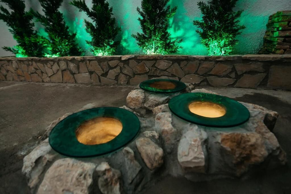 Drie groene schalen op stenen voetstukken in een donker gelegen buitenruimte, met een stenen muur en verlichte bomen op de achtergrond.