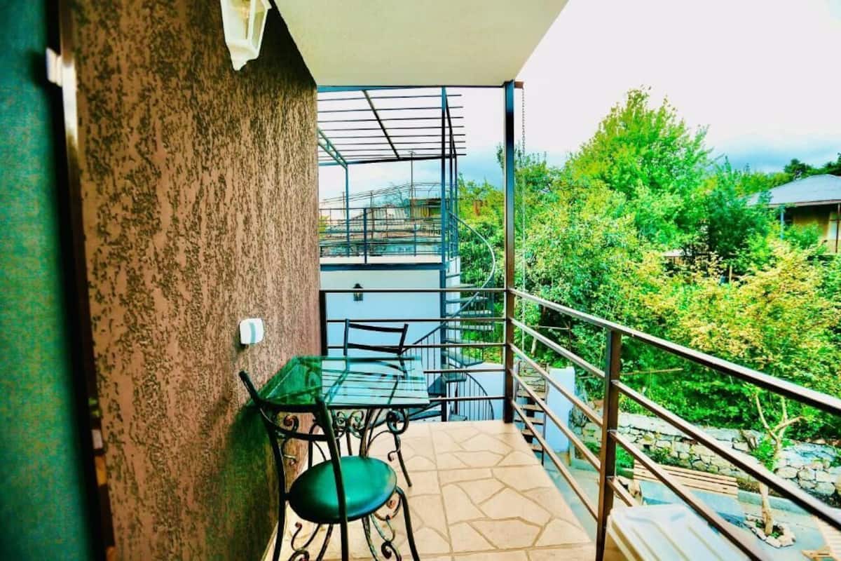 A balcony with a glass table and chairs, a metal railing, and a view of greenery and buildings.
