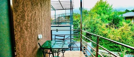 Un balcón con una mesa y sillas de vidrio, baranda de metal, y vistas a zonas verdes y edificios.