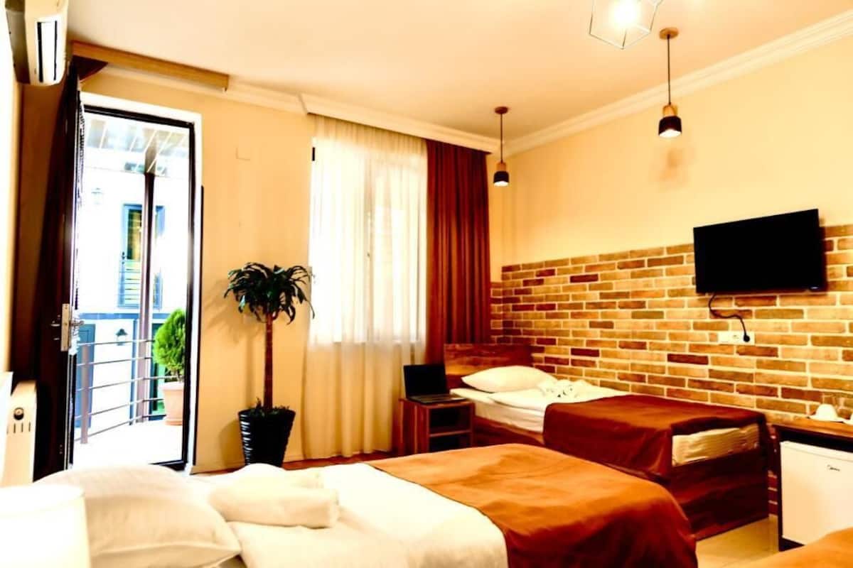 A hotel room with a brick wall, a TV mounted on the wall, a bed, a desk with a laptop, and a potted plant.