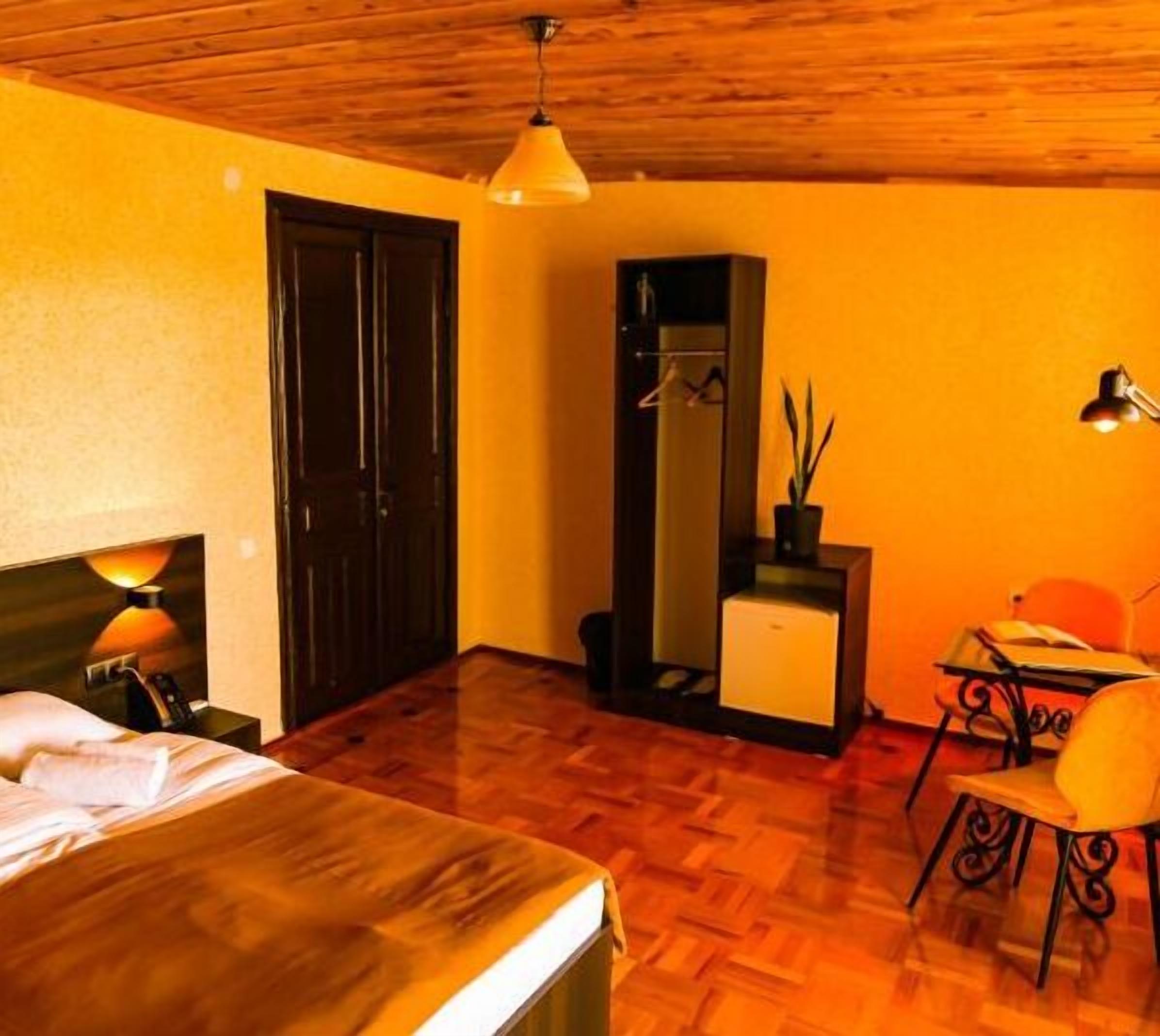 A hotel room with a wooden ceiling, a bed with white linens, a desk with a chair, a hanging light fixture, and a wardrobe.