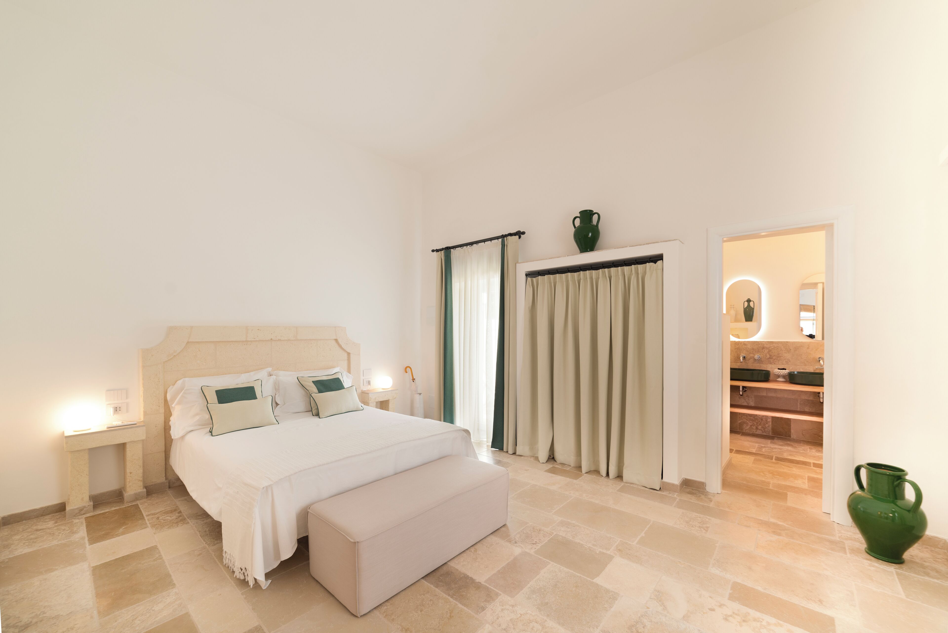 A bedroom with a bed, bedside tables, a nightstand, a bench, a bathroom visible through an open door, and a green vase on the floor.
