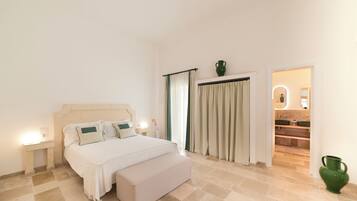 A bedroom with a bed, bedside tables, a nightstand, a bench, a bathroom visible through an open door, and a green vase on the floor.