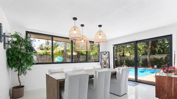 A modern dining area with a wooden table and white chairs, large windows, and a view of a swimming pool. - Tropical Beach House - Pool & Private Backyard - Close to everything (Boynton Beach)