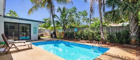 A swimming pool with lounge chairs, surrounded by palm trees and a wooden fence.