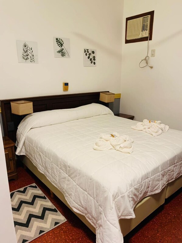 A neatly made bed with white bedding and towels, a wooden headboard, bedside tables, and a wall-mounted air vent.