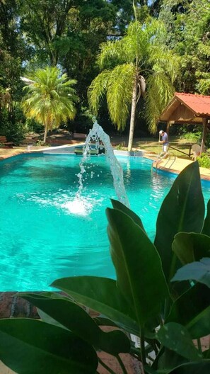 A swimming pool with clear blue water, surrounded by lush greenery and a wooden gazebo.