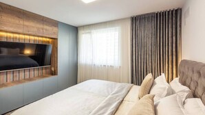 Ein modernes Schlafzimmer mit einem großen Bett, einem flachbildfernseher an einer Holzvertäfelung, einem Fenster mit transparenten Vorhängen und einem dunkel gefärbten Kopfteil.