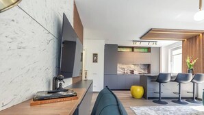 Ein modernes Wohnzimmer mit Flachbildfernseher, Couchtisch aus Holz und einer Couch. Es gibt eine Küchenzeile mit einer großen Kochinsel und Barhocker.
