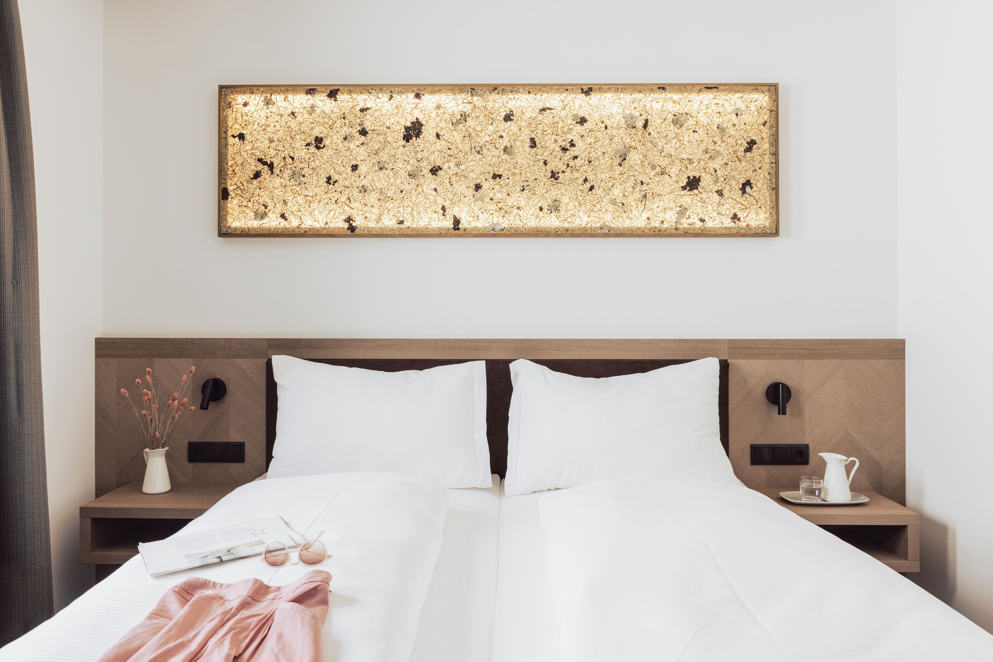 A double bed with white linens, two nightstands with a vase and a pitcher, and a wall-mounted artwork.