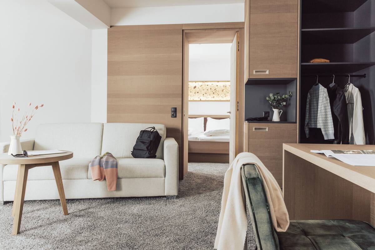 A modern hotel room with a sofa, a small round table, a desk, and a wardrobe.