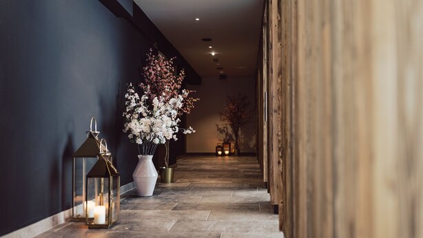 Un couloir avec un mur sombre, un panneau en bois, un vase blanc rempli de fleurs et une lanterne avec des bougies.
