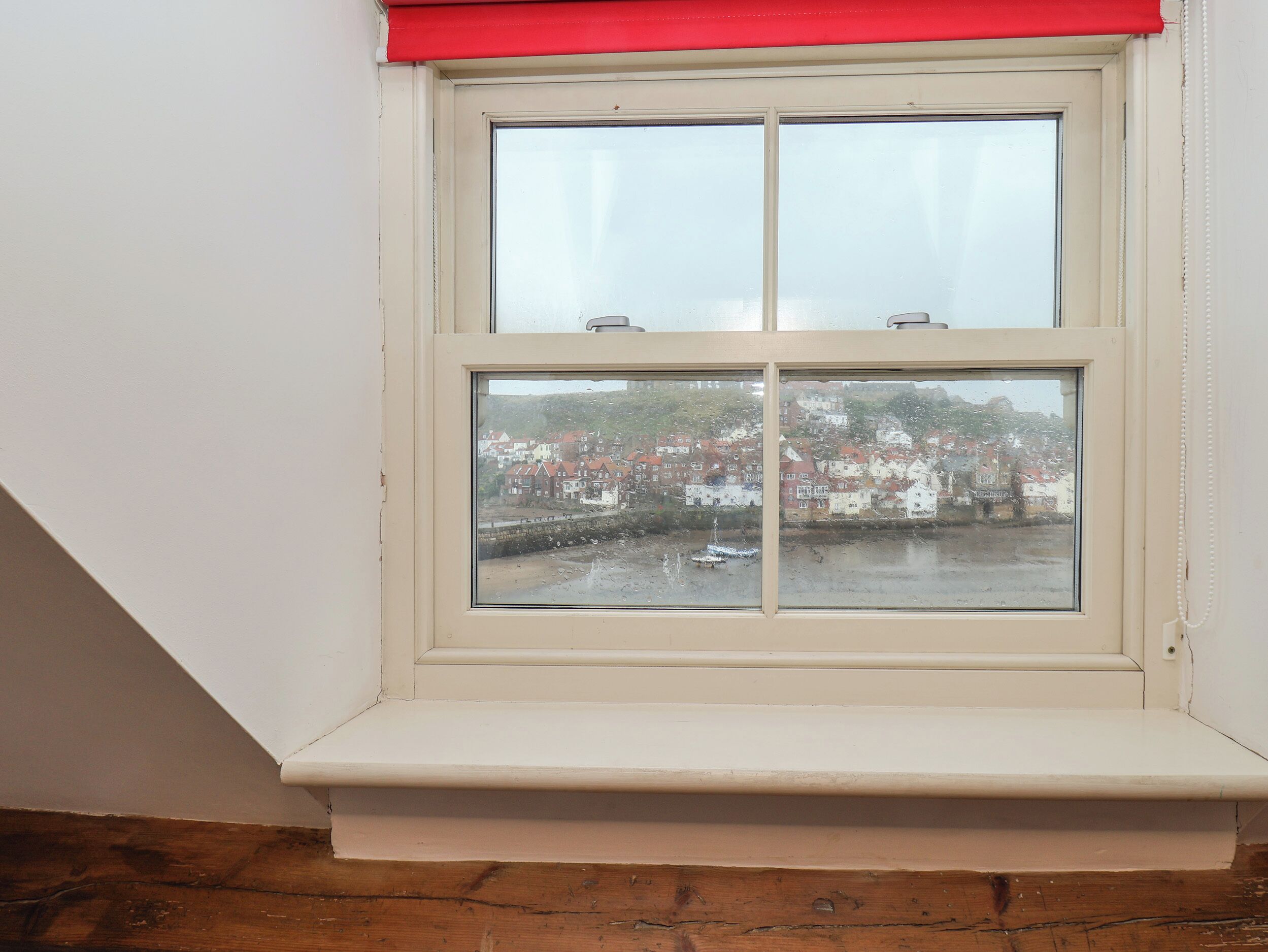 A window with a white frame and a red valance, overlooking a residential area with houses and a body of water.