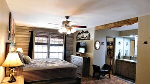 A bedroom with a bed, bedside lamps, a ceiling fan, a dresser, a TV, and a bathroom with a sink and mirror.