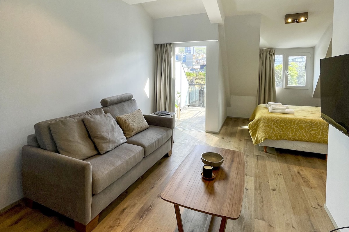 Ein modernes, helles Zimmer mit einer Couch, einem Couchtisch, einem Bett mit gelber Tagesdecke und einem Fernseher.