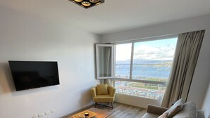 Un saló modern amb un televisor de pantalla plana, una taula de centre de fusta, un butaquito groc, un sofà beix i una finestra gran amb vista al mar i embarcacions.
