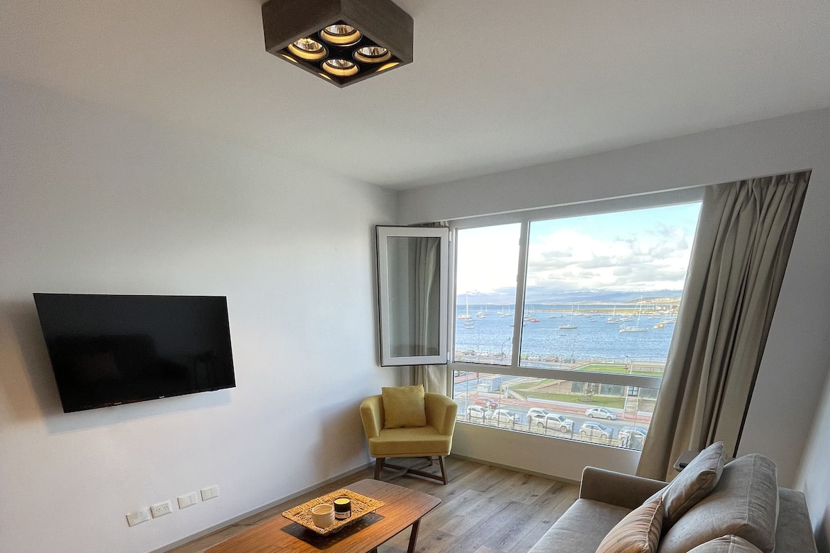 Ein modernes Wohnzimmer mit Flachbildfernseher, Couchtisch aus Holz, beiges Sofa, gelber Ohrensessel und einem großen Fenster mit Blick auf das Meer und Boote.