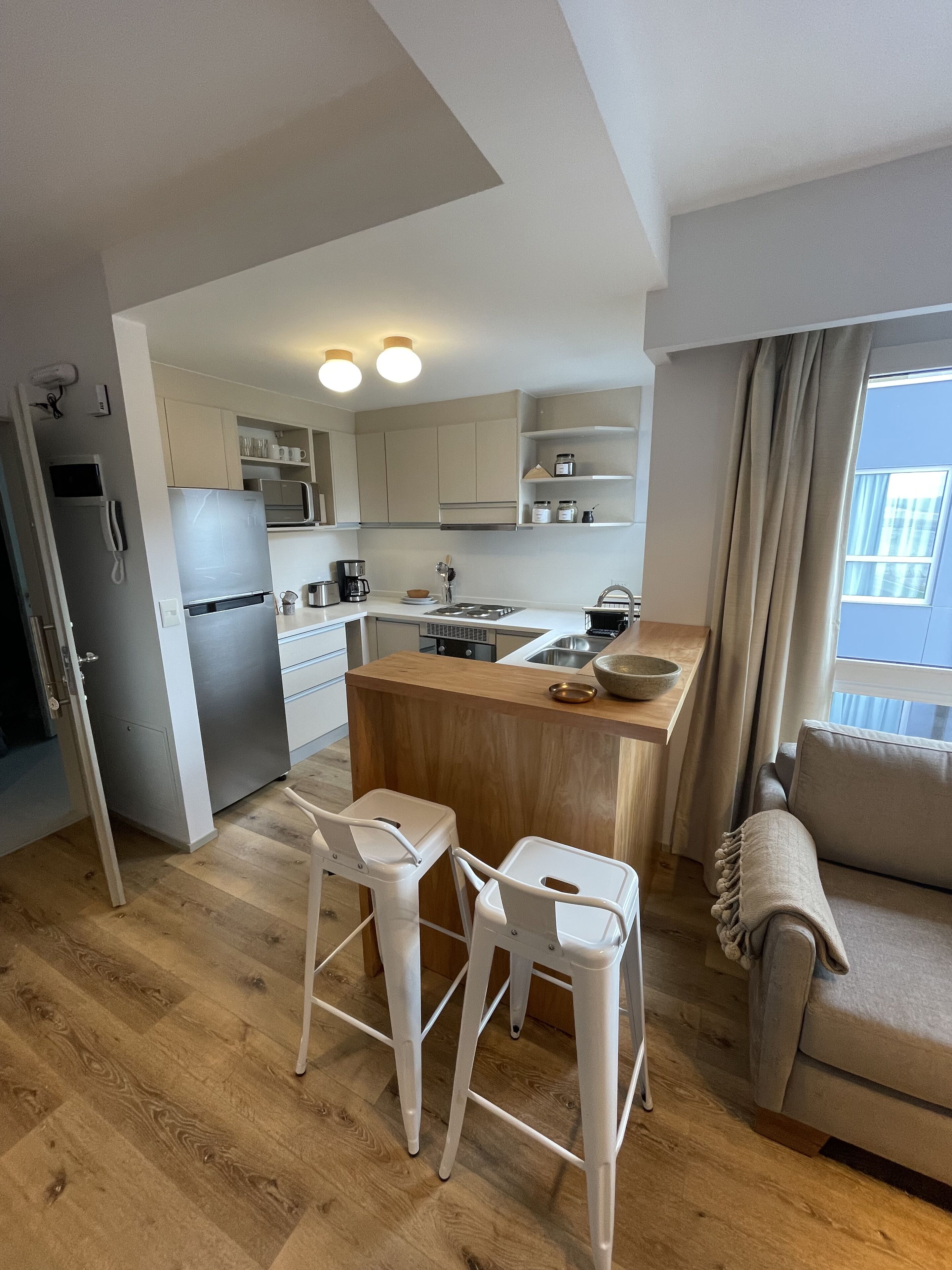 Ett modernt kök med vita skåp, en köksö i trä och en inbyggd ugn. Det finns två vita barstolar och en beige soffa.