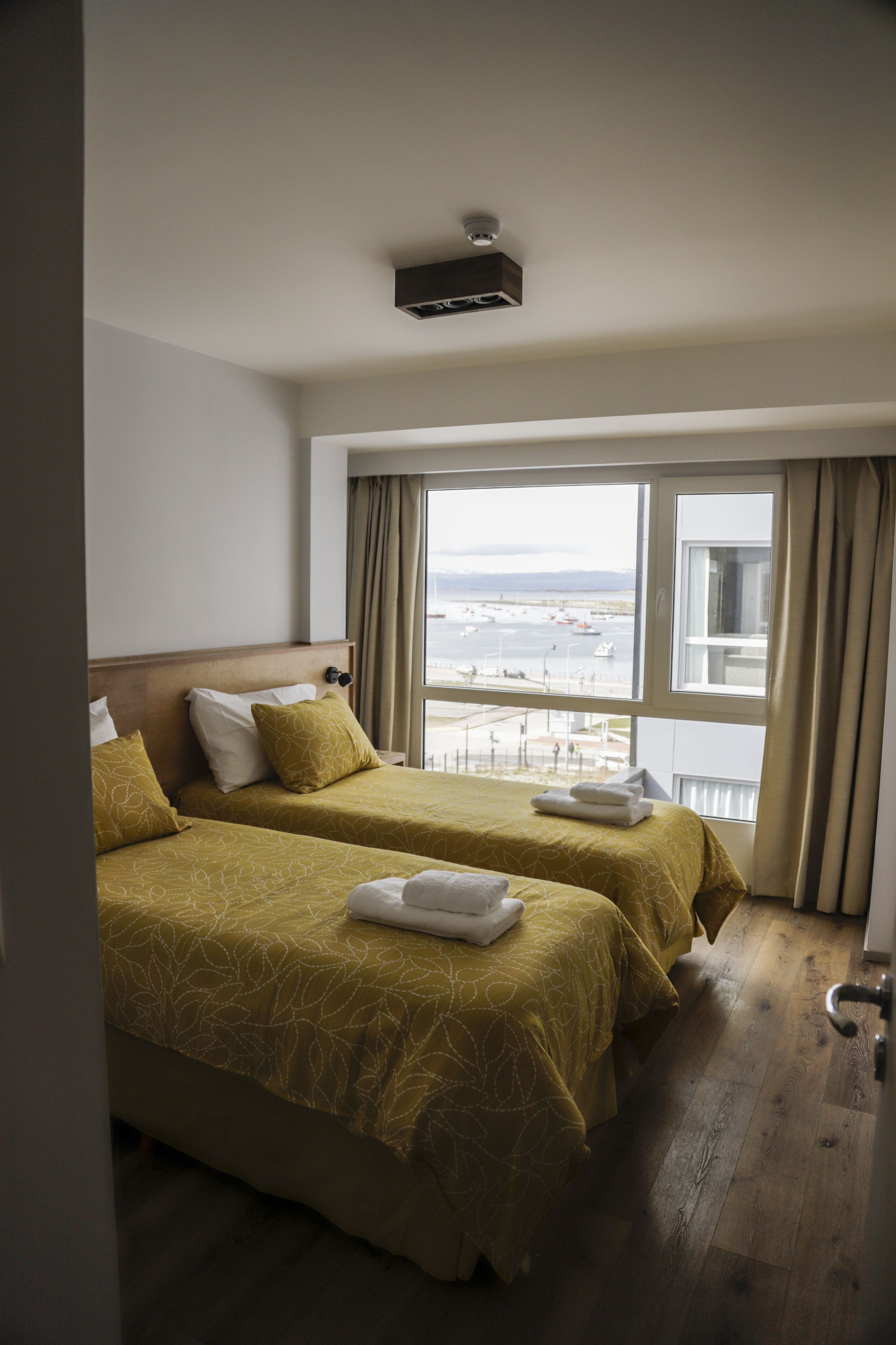 A hotel room with two beds, a large window with a view of the sea, and a ceiling-mounted air conditioning unit.