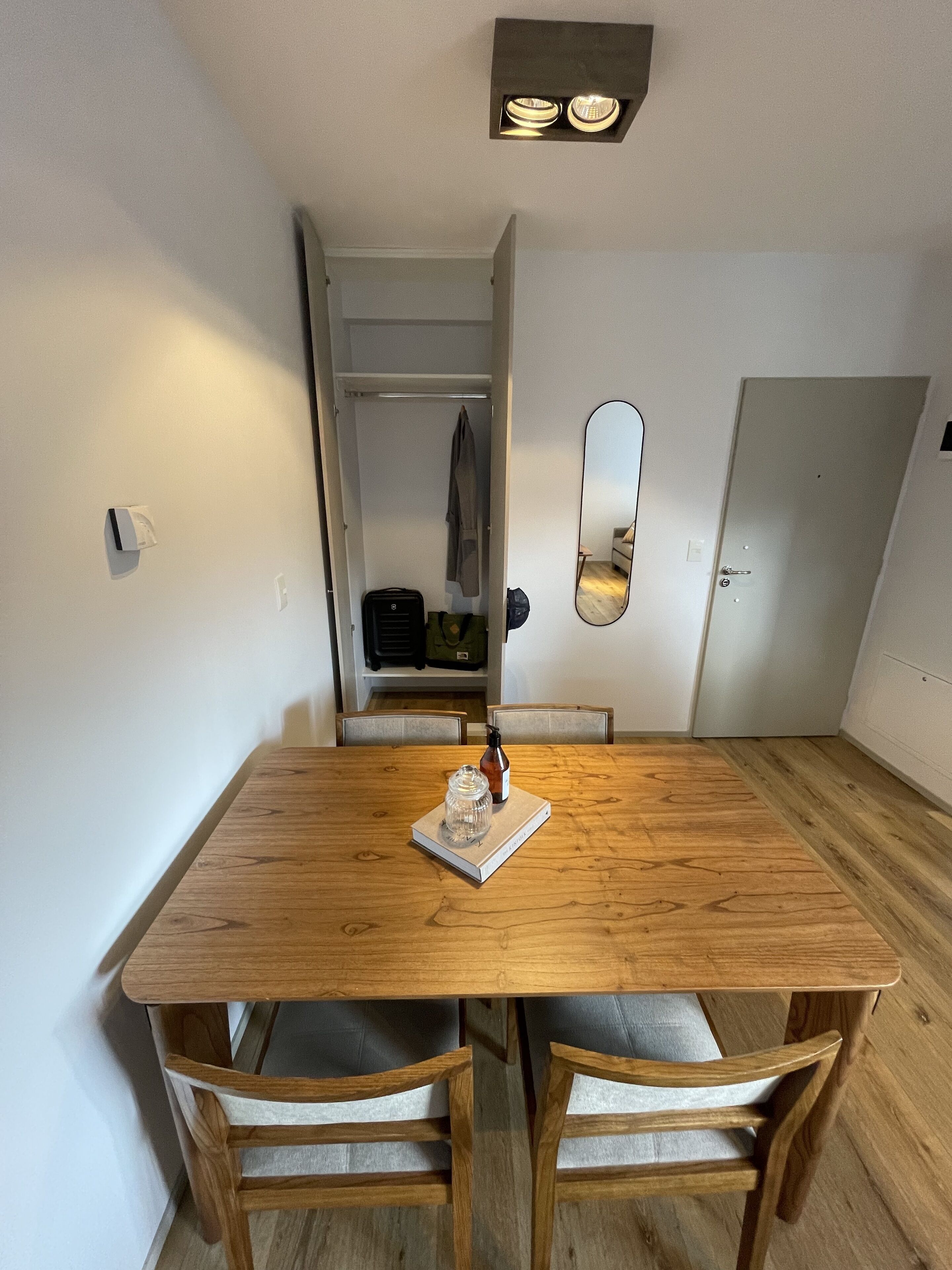 Una mesa de comedor de madera con dos sillas, un espejo pequeño y una lámpara colgante.