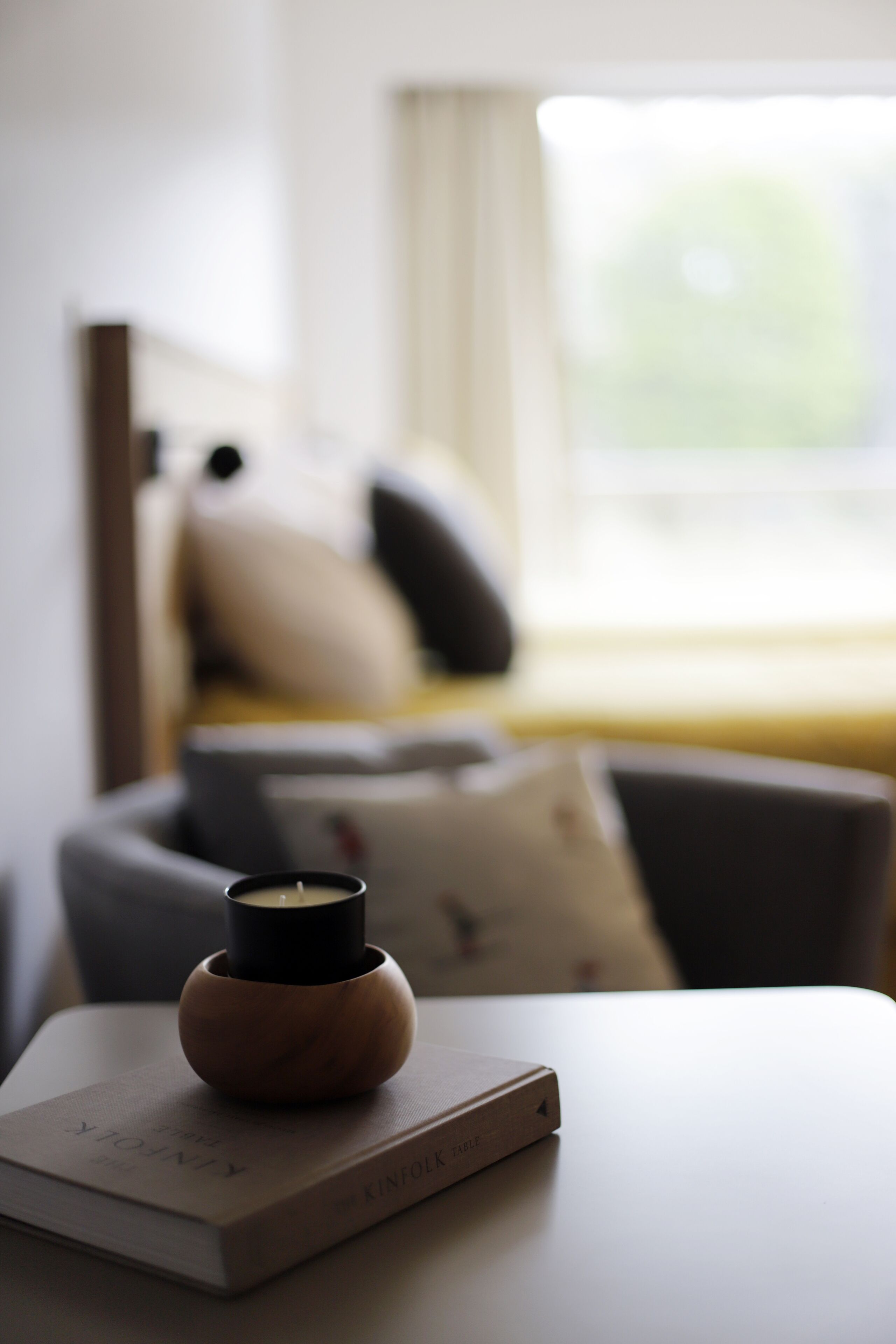 Une chambre avec un lit, une table de chevet sur laquelle se trouvent un livre et une bougie, et une fenêtre avec des rideaux.