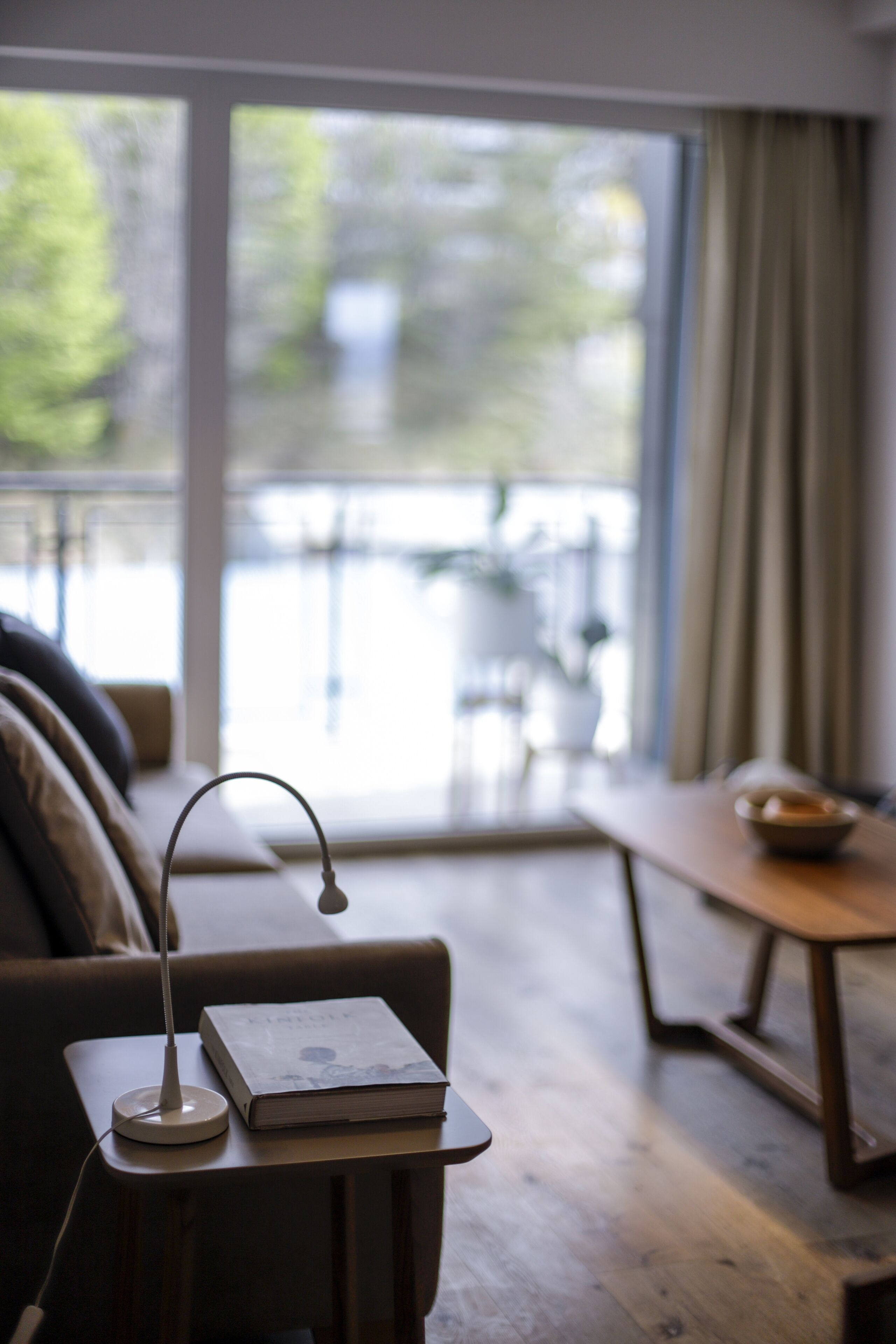 Ein gemütliches Wohnzimmer mit einem modernen Sofa, einem kleinen Tisch mit Lampe und Buch sowie Blick auf einen Balkon mit Gartenmöbeln.
