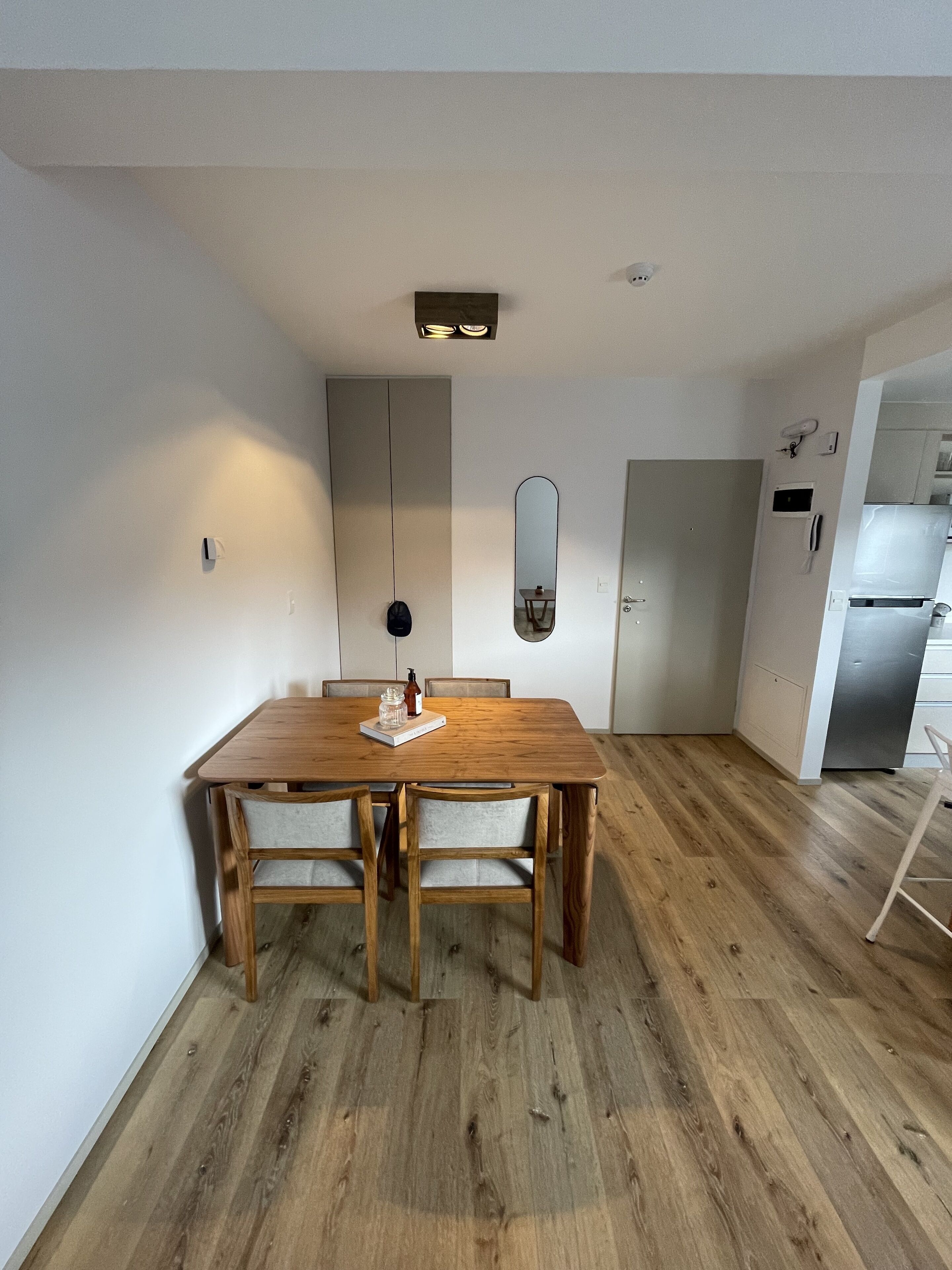 A modern dining area with a wooden table and chairs, a mirror on the wall, and a kitchen visible in the background.