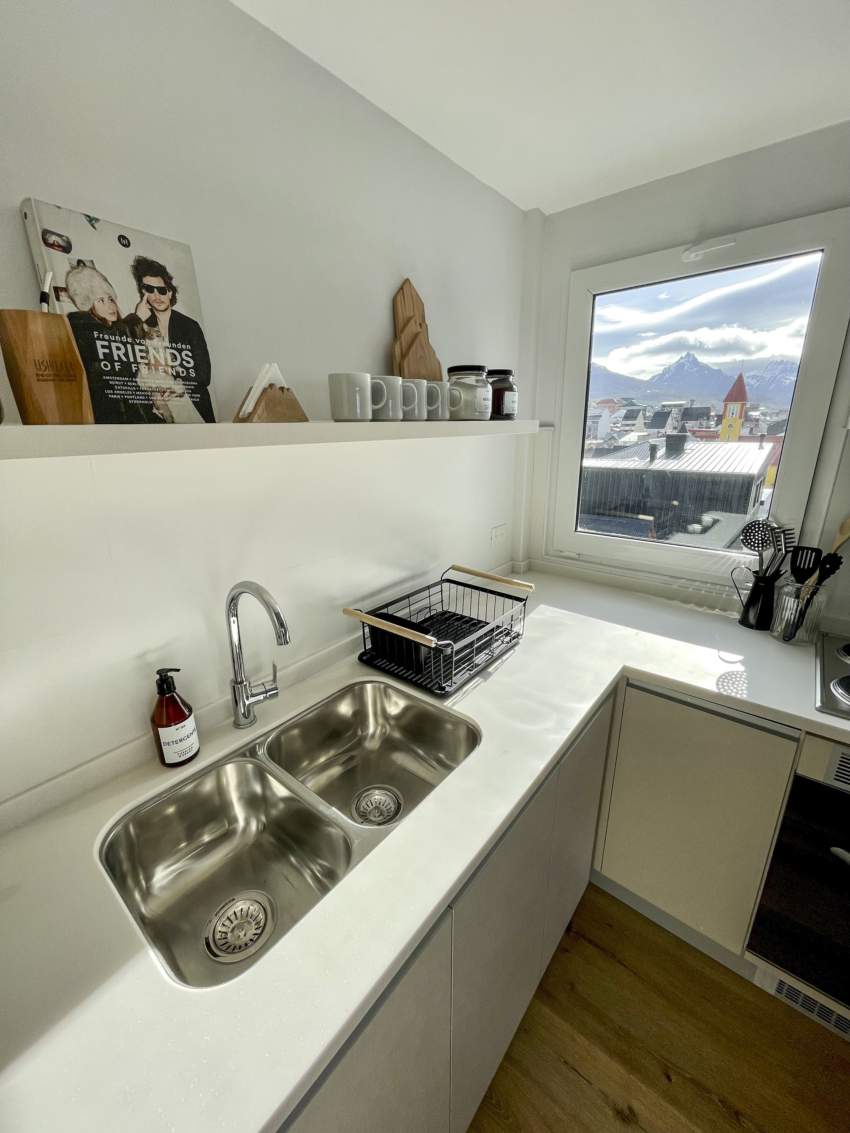 Une cuisine moderne dotée d’un évier à deux bacs, d’une fenêtre offrant une vue sur les montagnes et d’une étagère où sont rangés divers objets, dont un livre et des bocaux.
