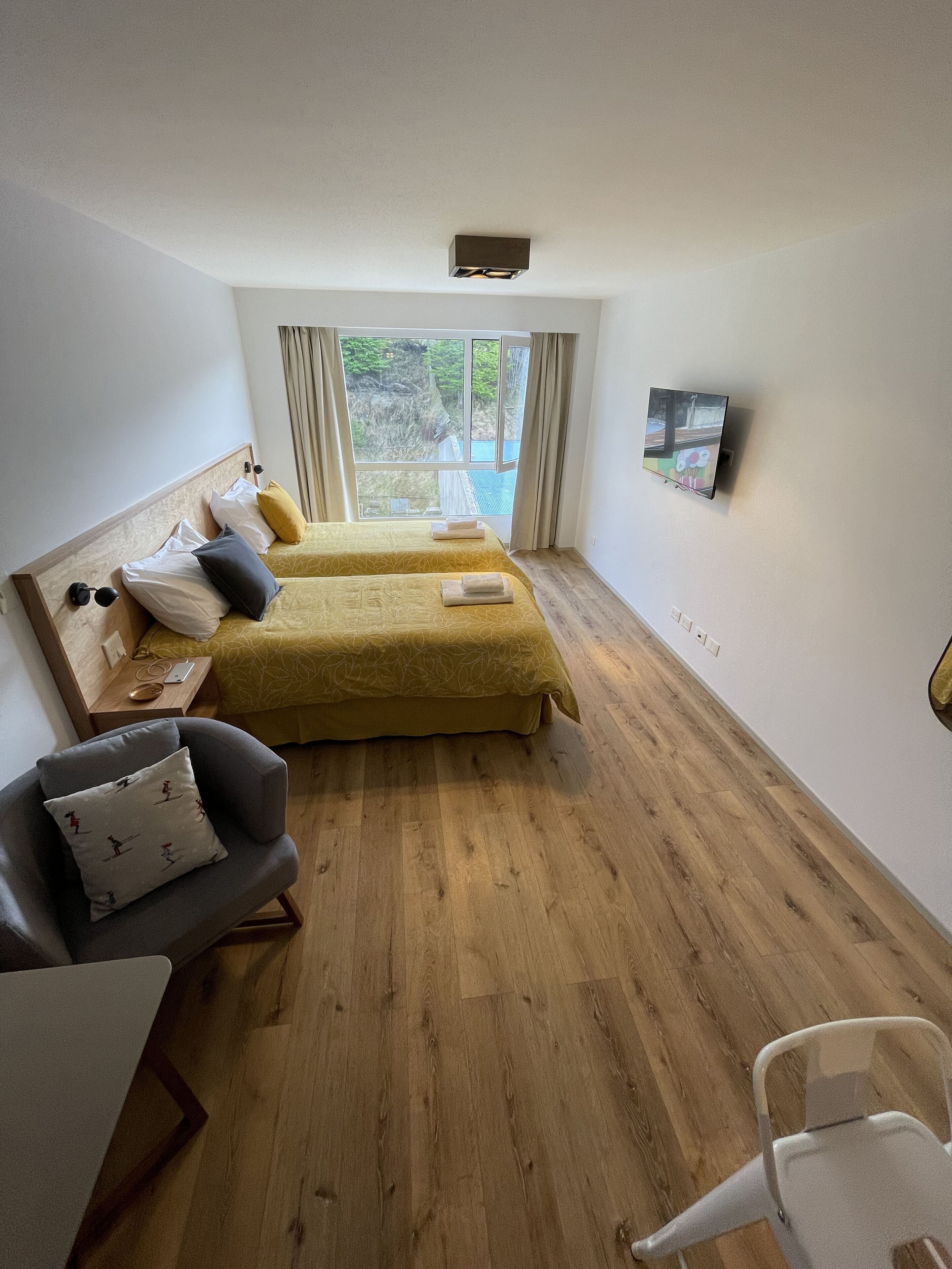 Habitación con dos camas, un escritorio y una silla. Cuenta con una ventana con cortinas, una lámpara en el techo y un televisor montado en la pared.
